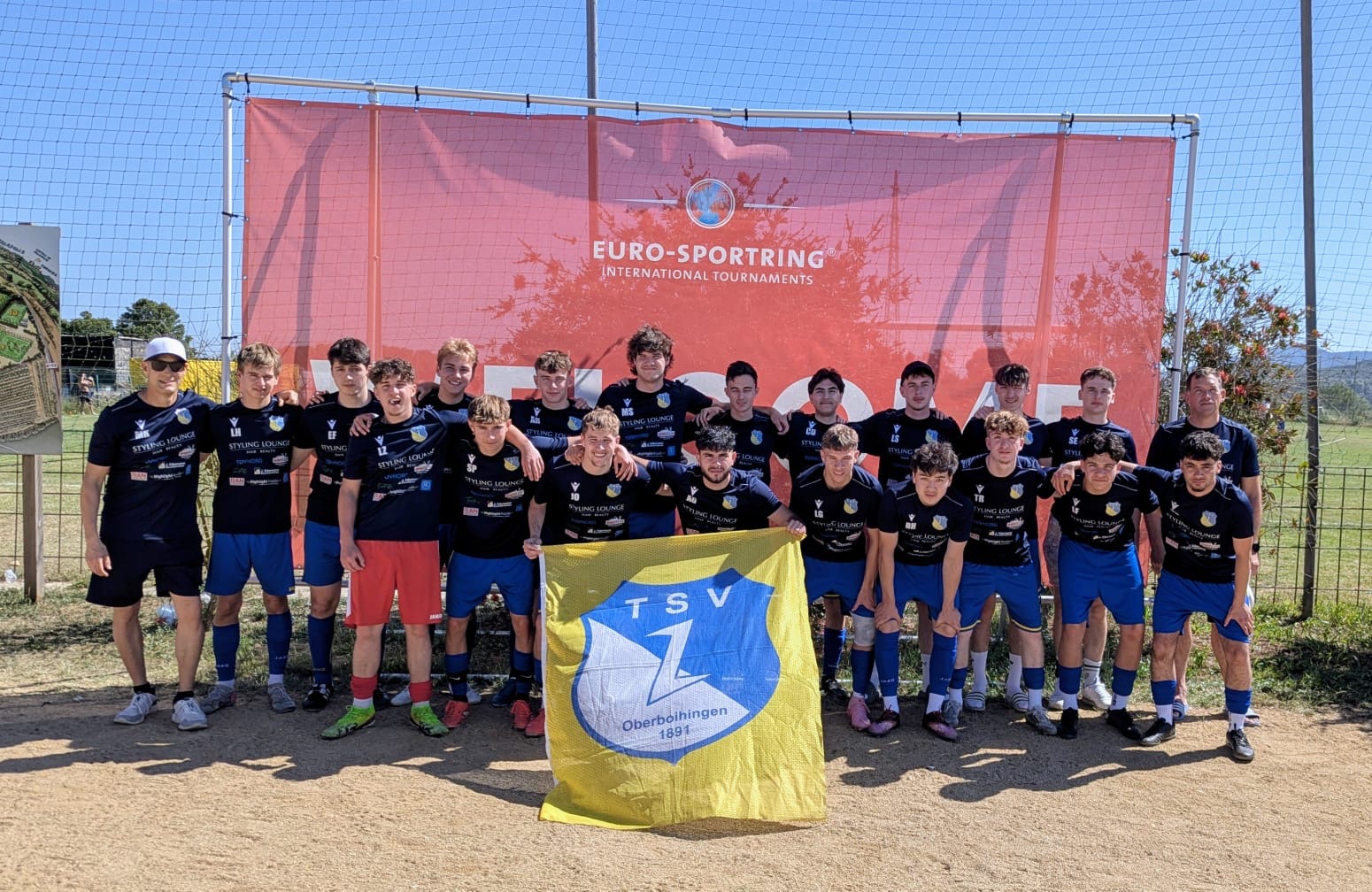 A-Junioren_Spanien_1 Spieler und Trainer des TSV Oberboihingen stehen als Mannschaftsgruppe vor einem Turnierbanner und halten die Vereinsflagge. Die Jugendfußballer tragen dunkle Trainingsshirts und blaue Hosen auf dem Gelände eines internationalen Fußballturniers.